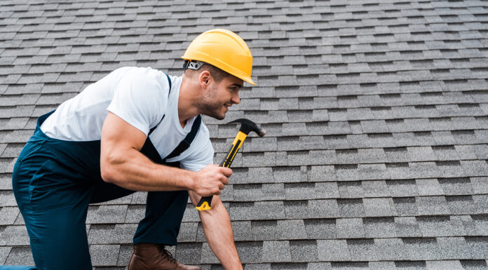 Why Regular Roof Maintenance Saves You Big Time handsome repairman in helmet holding hammer while repairing roof in house. Photo via adobe free stock