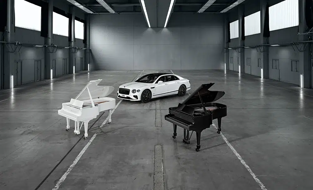 bentley-steinway-ultra-black-white-grand-pianos-collaboration.webp A Bentley Flying Spur Speed in Arctic White stands between Steinway’s Ultra Black and Ultra White grand pianos inside an industrial gallery, symbolizing the union of design, music, and motion.