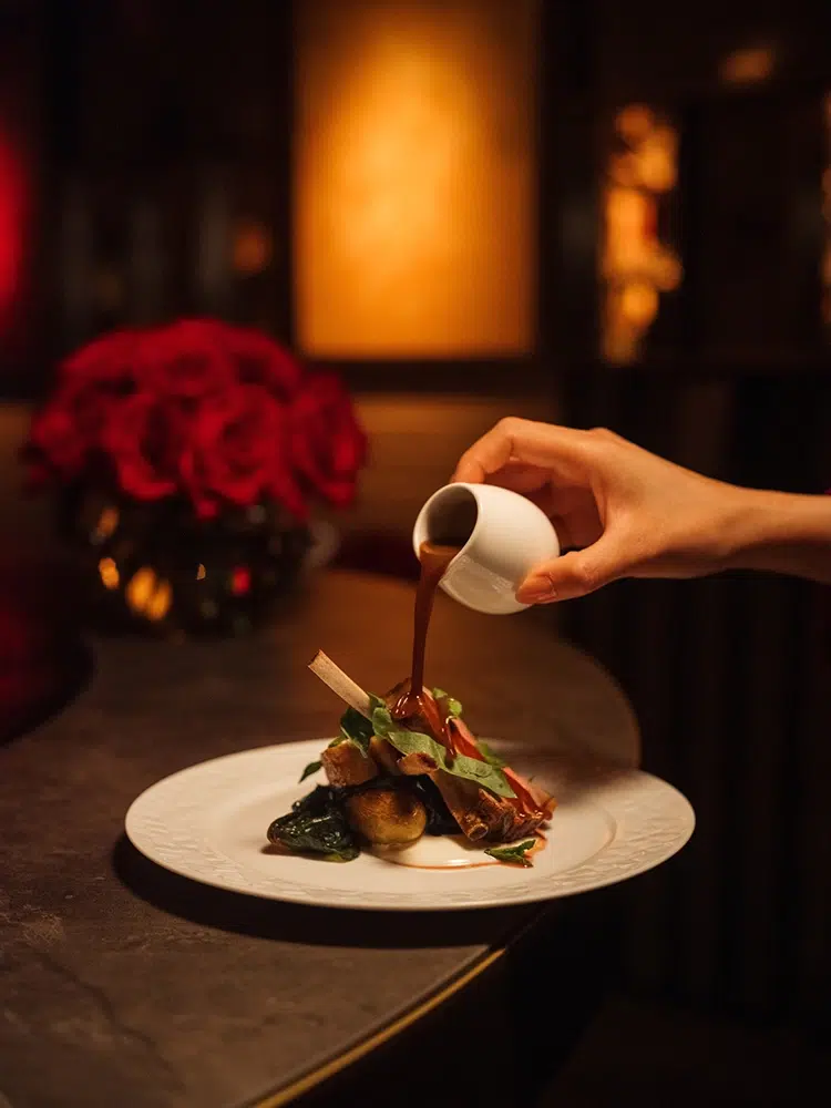 A Qatar Airways premium-cabin dish created by James Knappett receives its finishing sauce, reflecting the restaurant-inspired presentation of the airline’s new seasonal menu. James Knappett Qatar Airways Business Class dish with sauce presentation